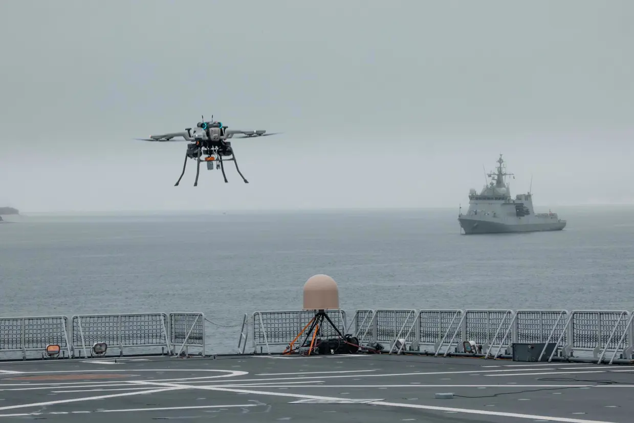 Een drone vliegt boven het dek van een marineschip. Op de achtergrond een marineschip op zee.