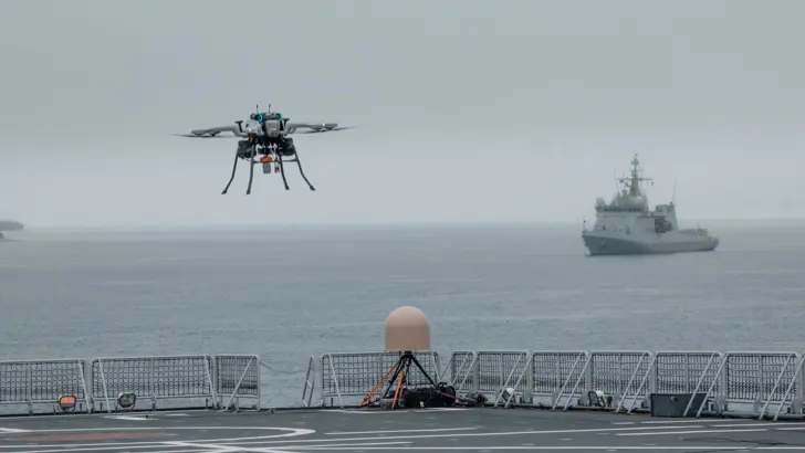 Een drone vliegt boven het dek van een marineschip. Op de achtergrond een marineschip op zee.