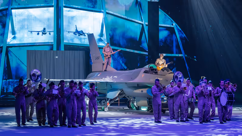 Op een blauw-paars verlicht podium bespelen muzikanten van de luchtmacht op en rondom een F-16 hun instrument.