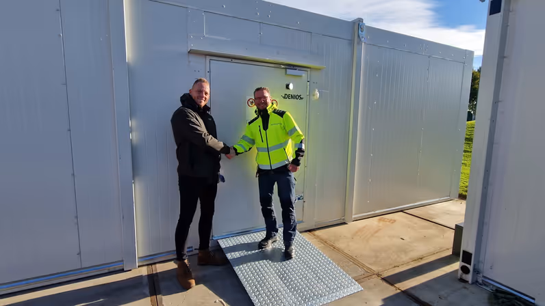 Glenn Afman in zwarte winterjas schudt de hand van een man in felgele werkjas. Ze staan buiten voor en tussen witte containers.
