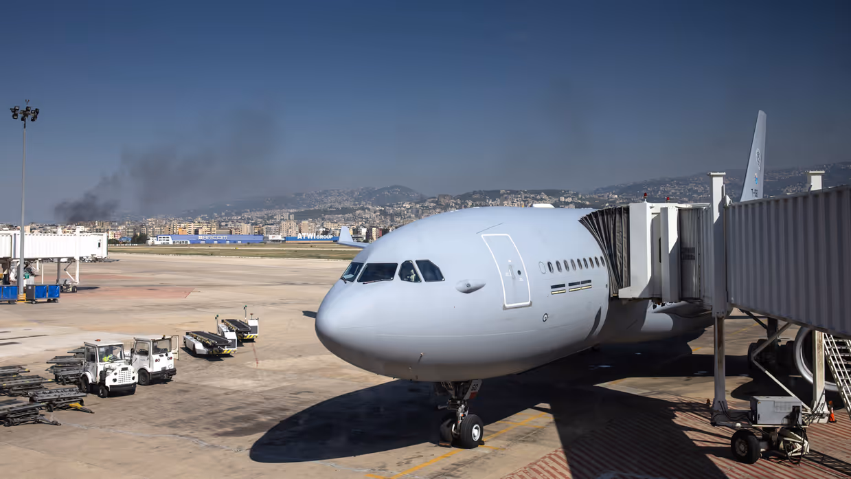 Een grijs, militair vliegtuig staat geparkeerd bij de gate. Op de achtergrond is een stad te zien waaruit rookwolken opstijgen.