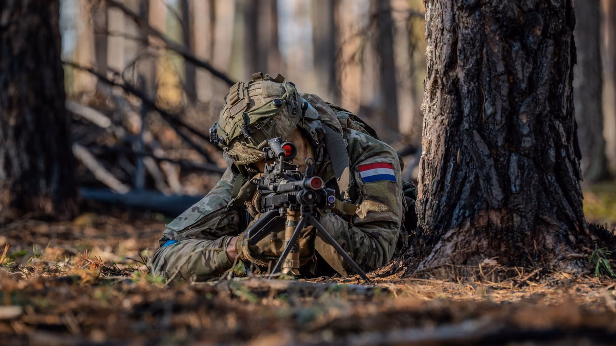 Militair met wapen ligt naast boom.