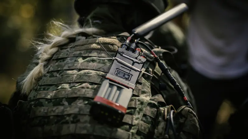 Een vrouwelijke militair vanaf de rug bezien. Ze heeft blond haar in een vlecht en draagt een helm. Op haar rug prijkt een radio met een zwarte antenne.&nbsp;