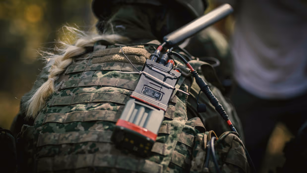 Een vrouwelijke militair vanaf de rug bezien. Ze heeft blond haar in een vlecht en draagt een helm. Op haar rug prijkt een radio met een zwarte antenne.&nbsp;