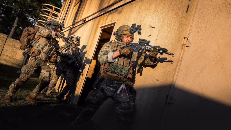 Een booskijkende militair gaat voorwaarts met een lang wapen langs zijn lichaam. Hij en de collega’s achter hem staan op het punt het gebouw rechts van hen binnen te vallen. 