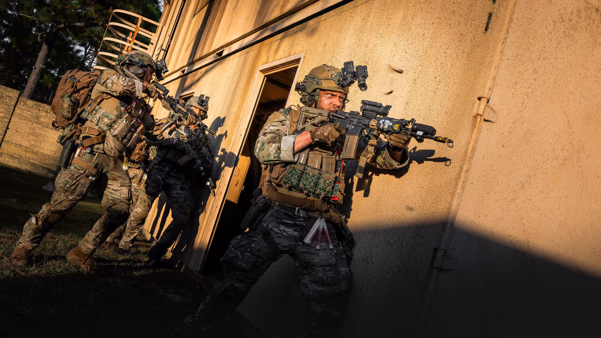 Een booskijkende militair gaat voorwaarts met een lang wapen langs zijn lichaam. Hij en de collega’s achter hem staan op het punt het gebouw rechts van hen binnen te vallen. 