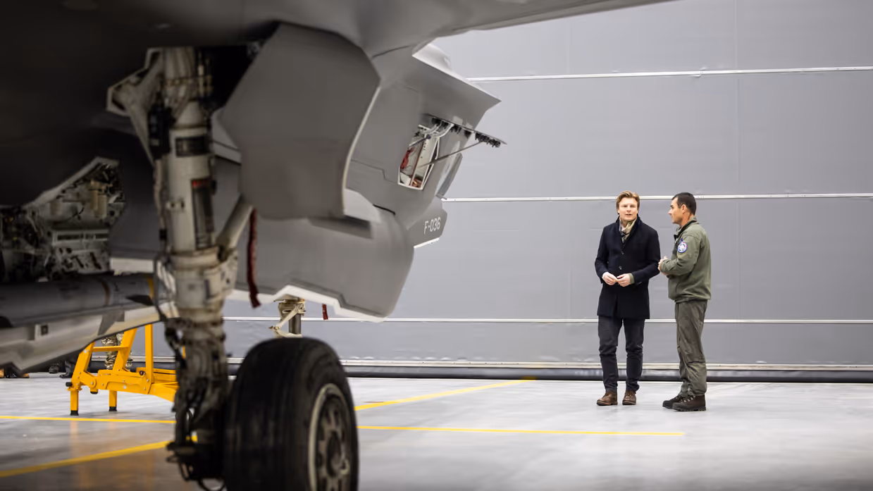Staatssecretaris Tuinman in groen pak staat de pers te woord. Minister Brekelmans praat onder een luchtmachter bij een F-35.