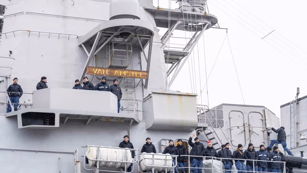 Militairen staan zwaaiend naast elkaar langs de reling van een marineschip.