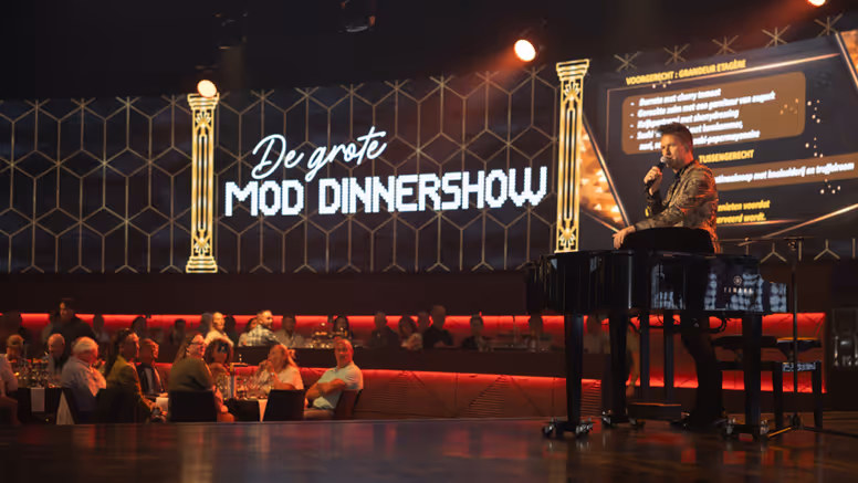 Een zaal vol mensen kijkt naar een man op een podium. Op de achtergrond is een billboard te zien met de tekst ‘De Grote MOD-dinnershow’.