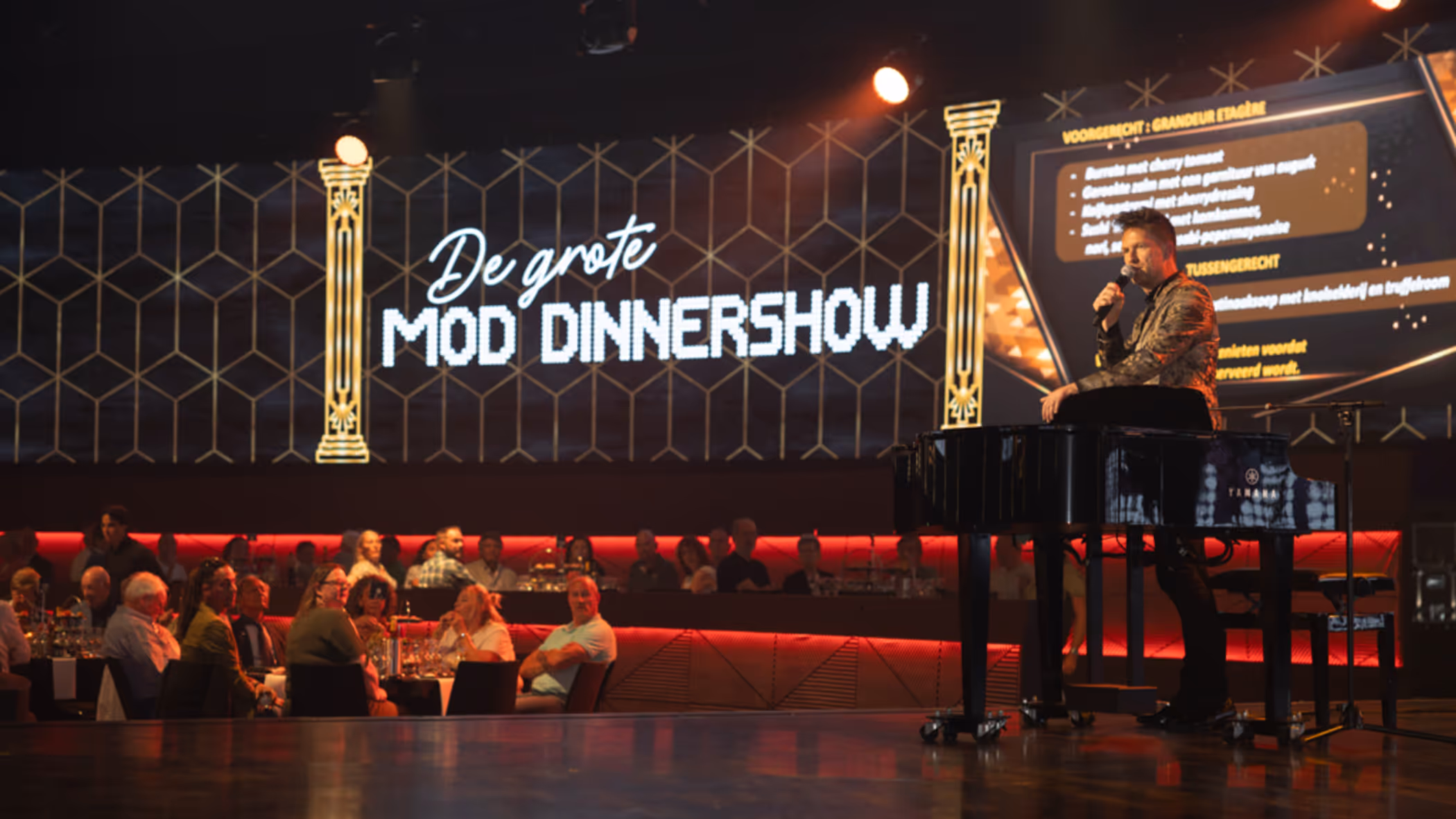 Een zaal vol mensen kijkt naar een man op een podium. Op de achtergrond is een billboard te zien met de tekst ‘De Grote MOD-dinnershow’.