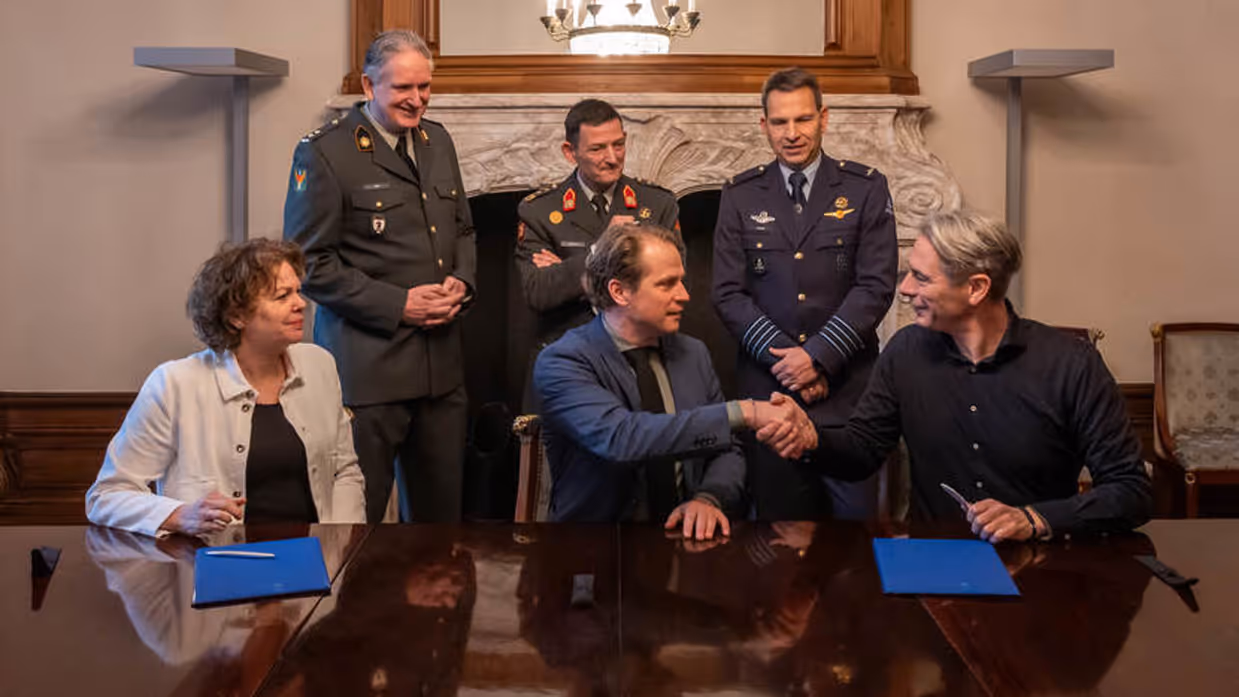 Drie mensen zitten aan een houten tafel. De rechter twee geven elkaar een hand. Achter hen staan drie militairen.