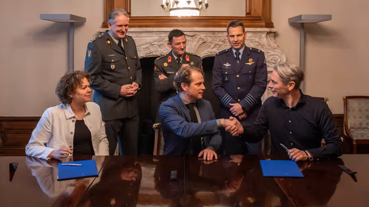 Drie mensen zitten aan een houten tafel. De rechter twee geven elkaar een hand. Achter hen staan drie militairen.
