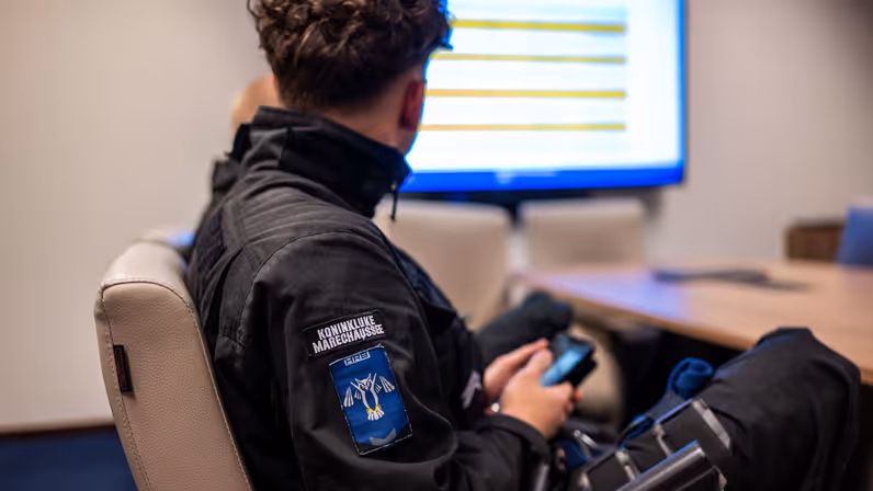 Een cursist van de Koninklijke Marechaussee volgt een briefing terwijl routes en aandachtspunten op een scherm worden besproken.