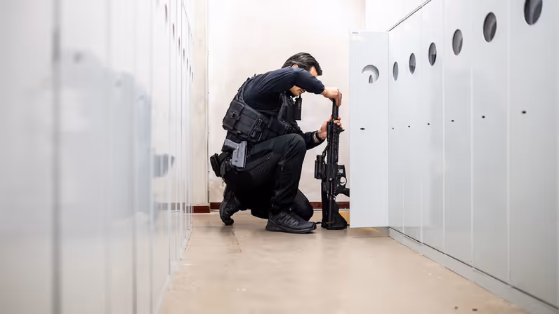 Een marechaussee knielt in een gang tussen kluisjes terwijl hij een lang wapen veilig voorbereidt voorafgaand aan de oefening. 