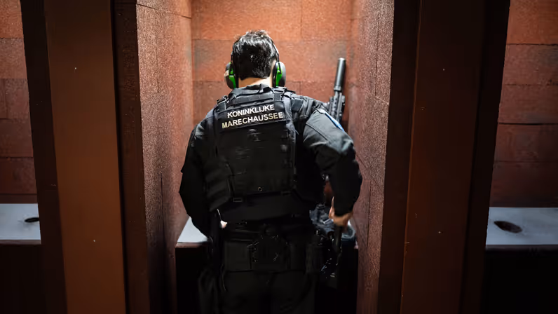 Een marechaussee staat in een ontlaad- en laadvoorziening met gehoorbescherming terwijl hij een lang wapen gereedmaakt.