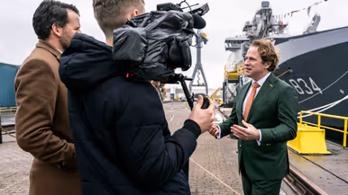 Staatssecretaris Tuinman in groen pak staat de pers te woord. Minister Brekelmans praat onder een luchtmachter bij een F-35.