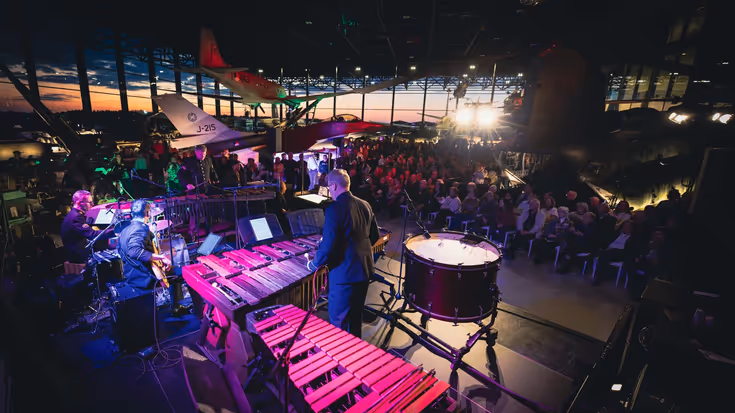 Militair orkest in sfeervol verlichte ruimte speelt muziek voor een publiek dat tussen vliegtuigen zit.