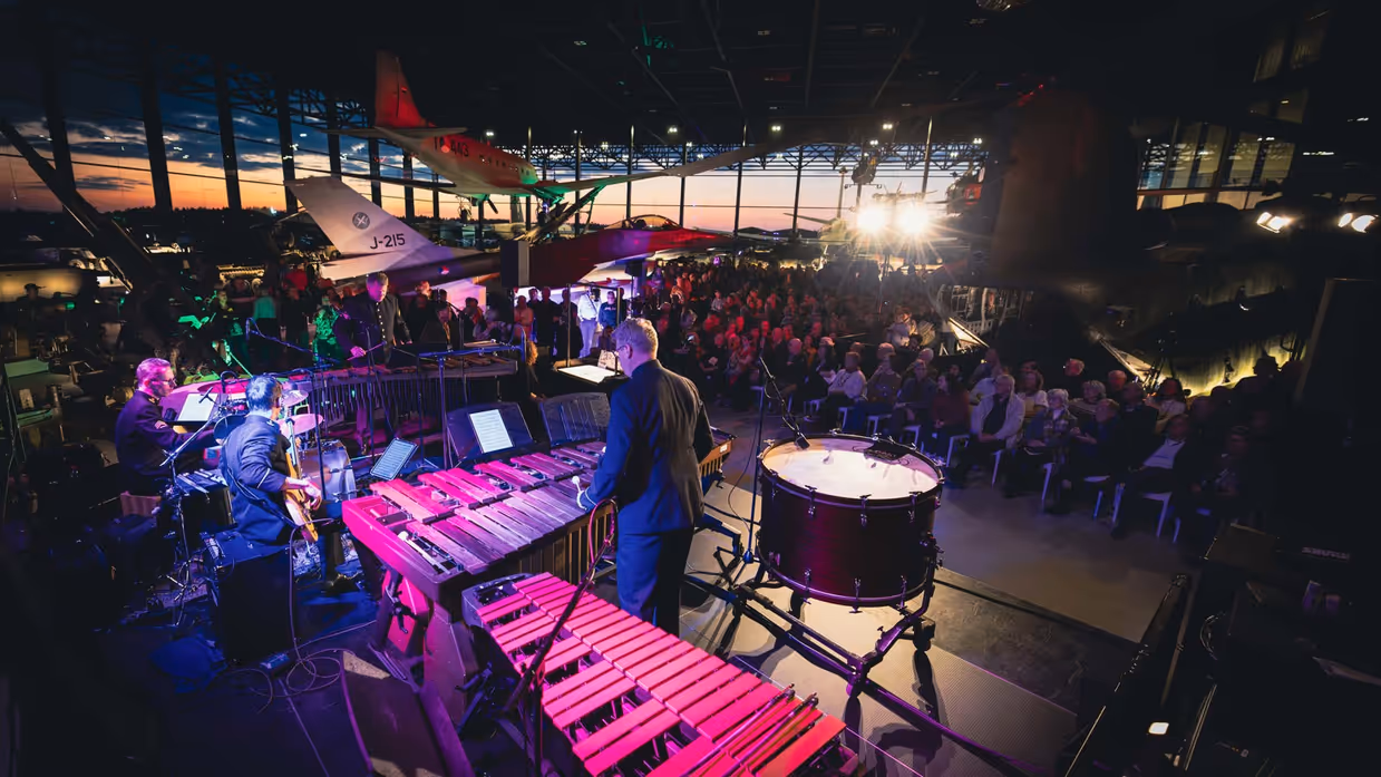 Militair orkest in sfeervol verlichte ruimte speelt muziek voor een publiek dat tussen vliegtuigen zit.