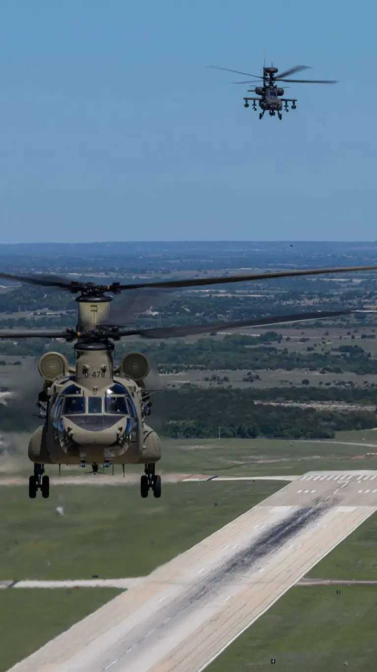 Een Chinook en twee Apaches vliegend boven een startbaan met op de achtergrond een blauwe lucht en weids uitzicht over vlak platteland.