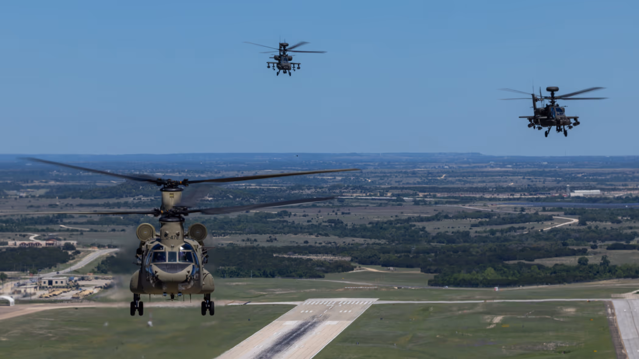 Een Chinook en twee Apaches vliegend boven een startbaan met op de achtergrond een blauwe lucht en weids uitzicht over vlak platteland.