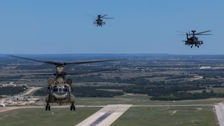 Een Chinook en twee Apaches vliegend boven een startbaan met op de achtergrond een blauwe lucht en weids uitzicht over vlak platteland.