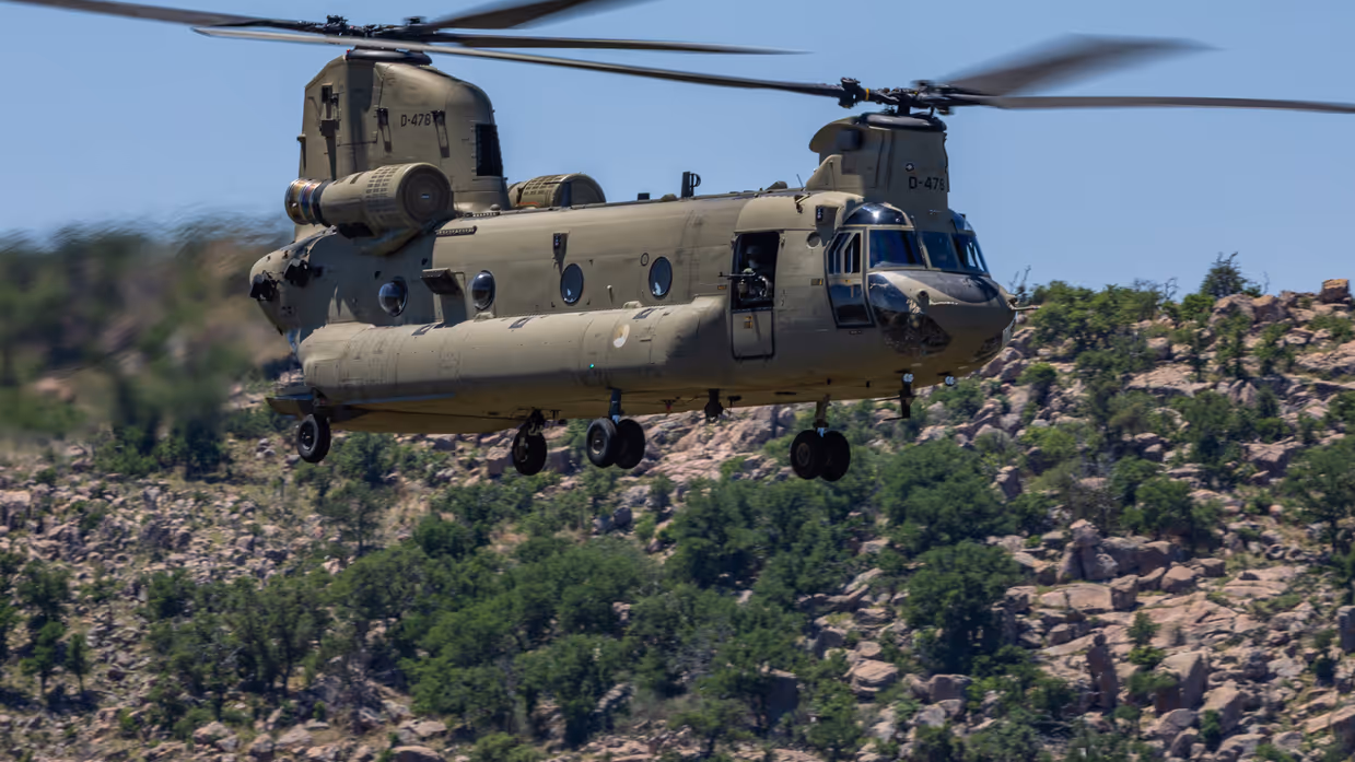 Een laagvliegende Chinook boven een heuvel met rotsblokken en begroeiing.