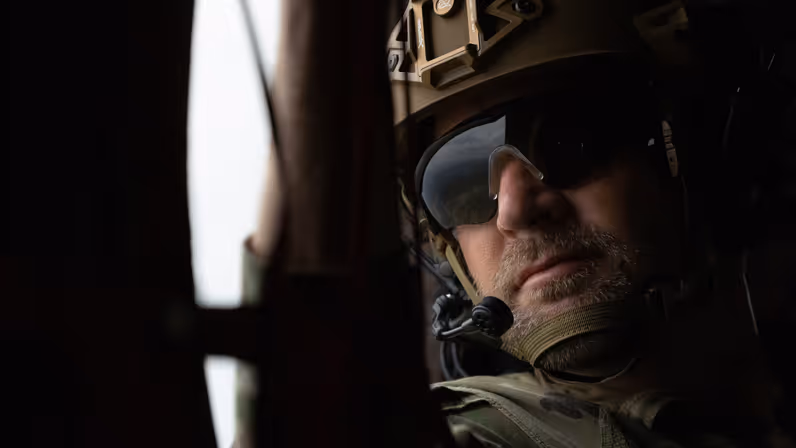 Close-up van militair met zonnebril en helm inclusief microfoontje voor de mond die uit een helikopterraam kijkt.