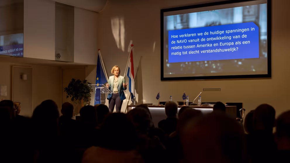 Vrouw spreekt vanaf podium volle zaal toe tijdens presentatie.