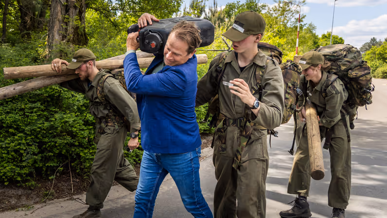 Tuinman in blauw jasje sjouwt een grote jerrycan op zijn schouders. 