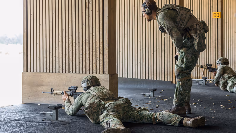 Een militair ligt op zijn buik met een wapen voor zijn gezicht op de schietbaan. Een andere militair staat achter hem en geeft hem instructies.&nbsp;