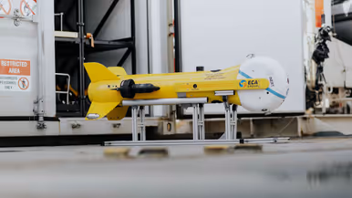 Links een grijs bootje met achterop verschillende drones. Dit vaart langs een groot vrachtschip. Daarnaast een gele, raketvormige drone met voorop een witte bol. Deze is ook te zien achterop het varende bootje.