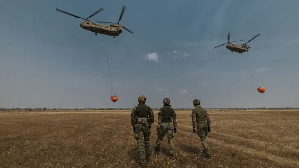 Militairen op de rug gezien. In de lucht 2 helikopters met waterzakken.