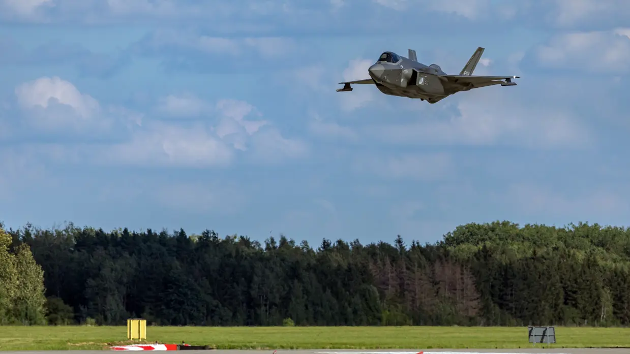 Een vliegende F-35, net boven de landingsbaan. Onder op de foto een bosrand, daarboven een blauwe lucht.