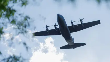 Een C-130 vliegt laag over. Links staan bomen. 