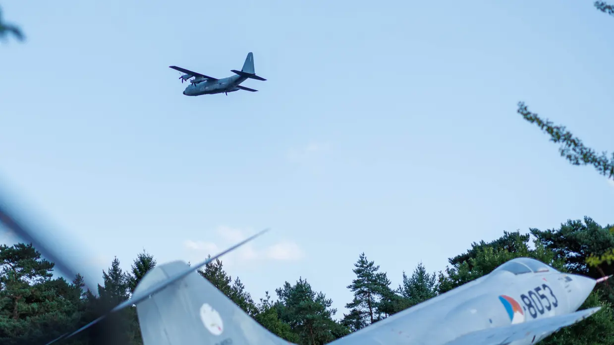 Een Italiaanse C-130 vliegt over een F-104G Starfighter die op de grond staat.  