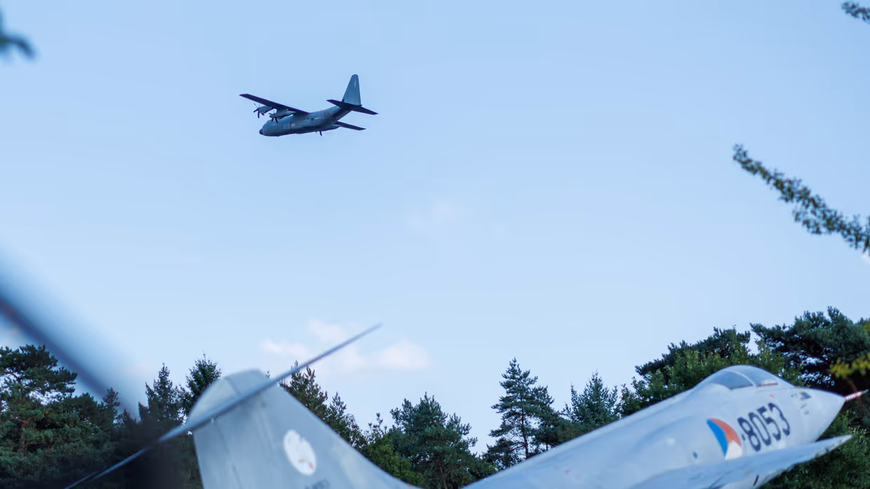 Een Italiaanse C-130 vliegt over een F-104G Starfighter die op de grond staat.  