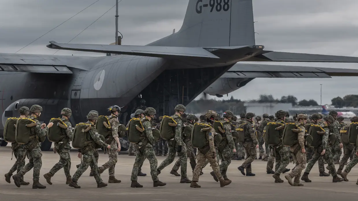 Hercules met op de voorgrond militairen die met een parachute op hun rug lopen. 