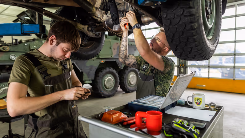 Een man in een groene overall sleutels aan een klein onderdeel in zijn handen. Een ander sleutels boven zijn hoofd aan de onderkant van een voertuig.&nbsp;