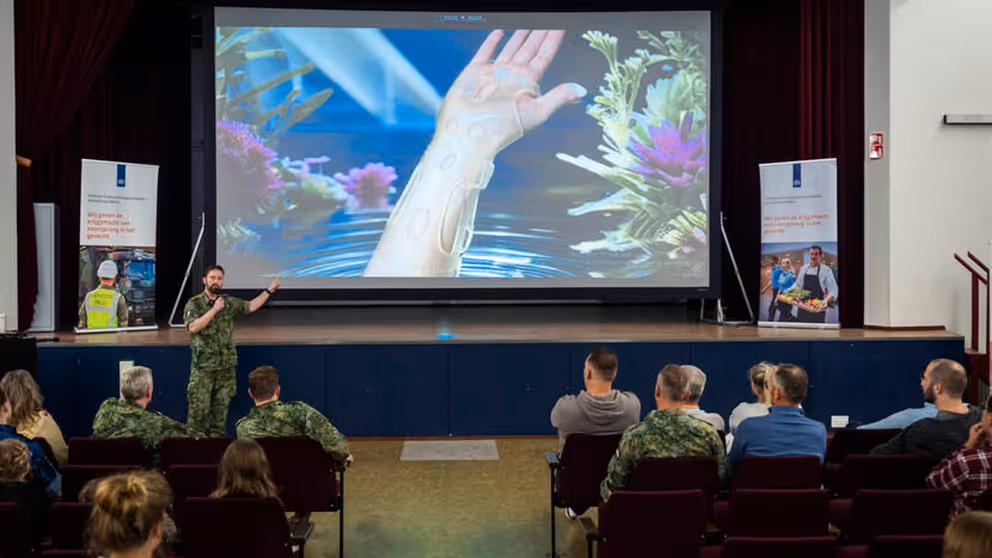 Spreker traumachirurg kolonel Vincent Stirler wijst naar een afbeelding op een scherm van een spalk die een patient om de pols heeft.