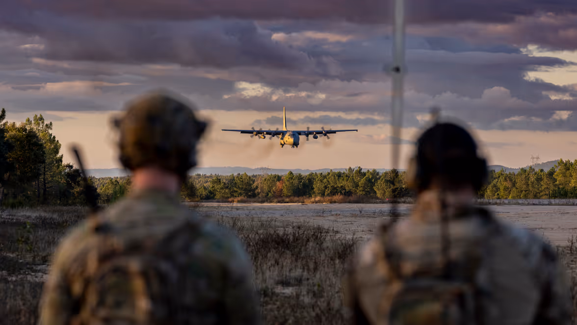 Een C-130 Hercules nadert een open vlakte, op de voorgrond twee militairen die vanuit de bosrand toekijken.