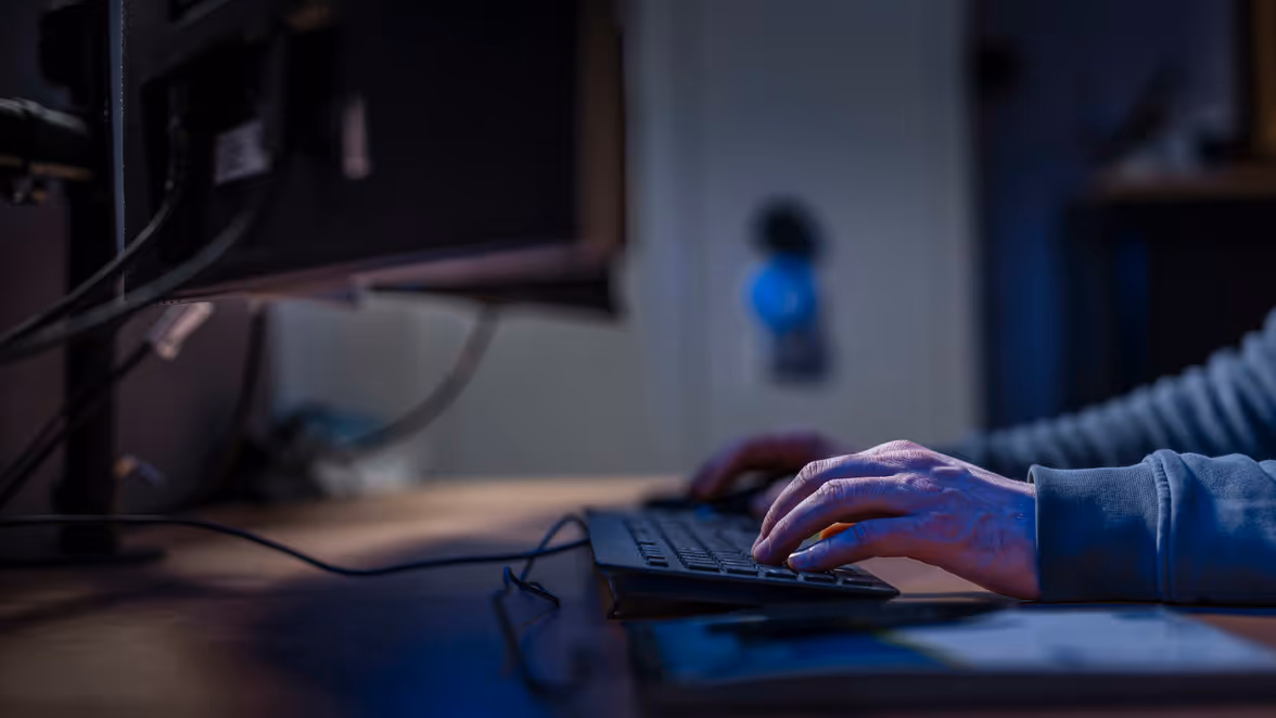 Close-up van handen die typen op een toetsenbord in een halfdonkere kantooromgeving met computerkabels en beeldschermen op de achtergrond.