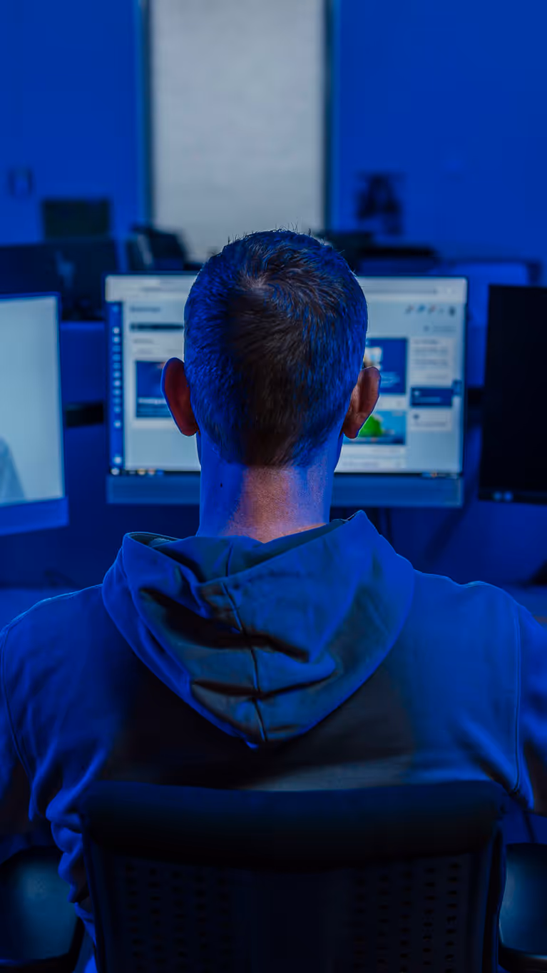 Professionele werkomgeving met een persoon aan een bureau, kijkend naar meerdere computerschermen in een blauw verlichte ruimte. Ook een silhouet van een gemaskerde figuur op één van de schermen.