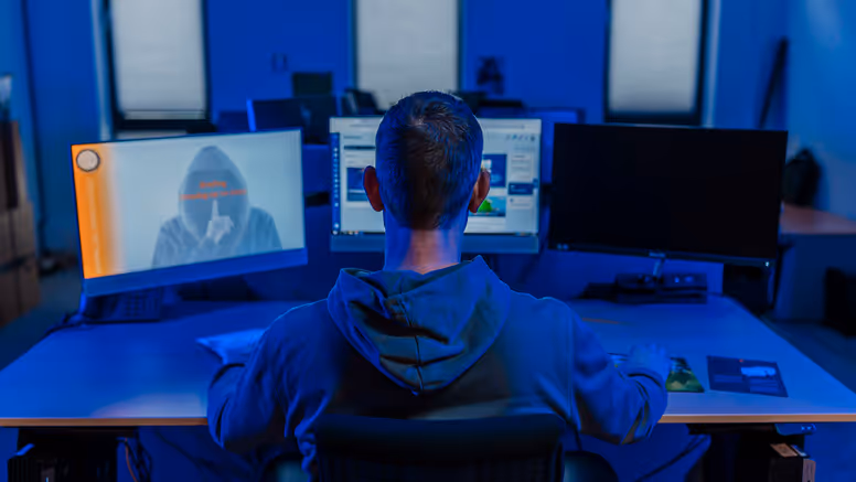 Professionele werkomgeving met een persoon aan een bureau, kijkend naar meerdere computerschermen in een blauw verlichte ruimte. Ook een silhouet van een gemaskerde figuur op één van de schermen.