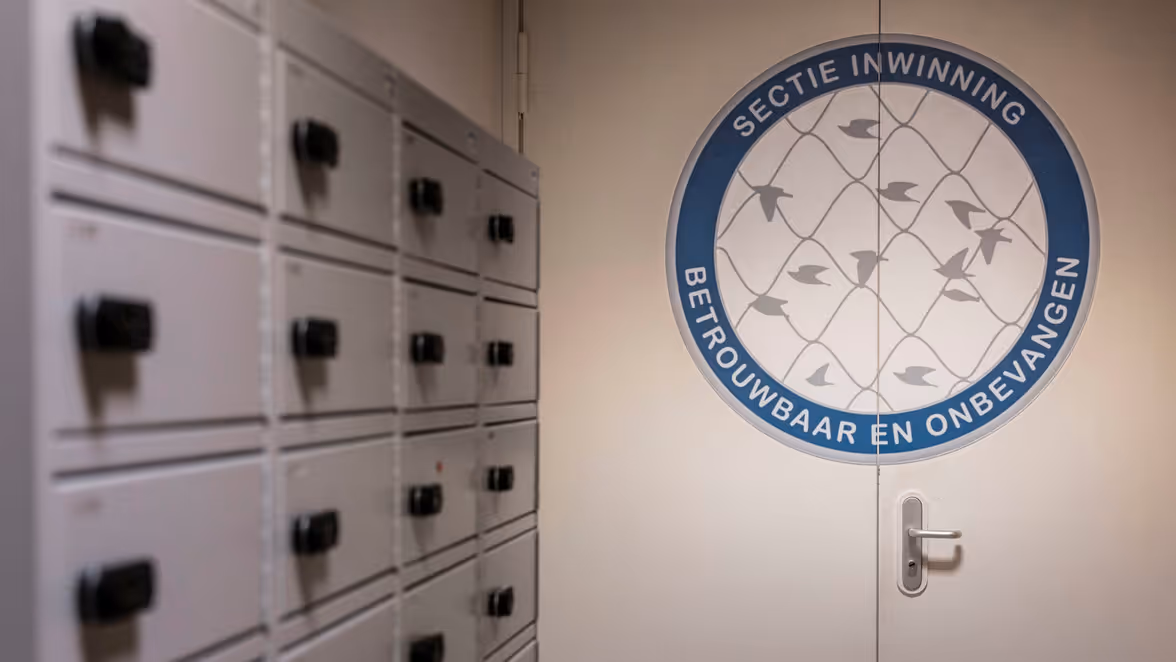 Gang met lockers naast een deur met een groot rond embleem waarop staat “Sectie Inwinning. Betrouwbaar en Onbevangen” en een patroon van vliegende vogels.