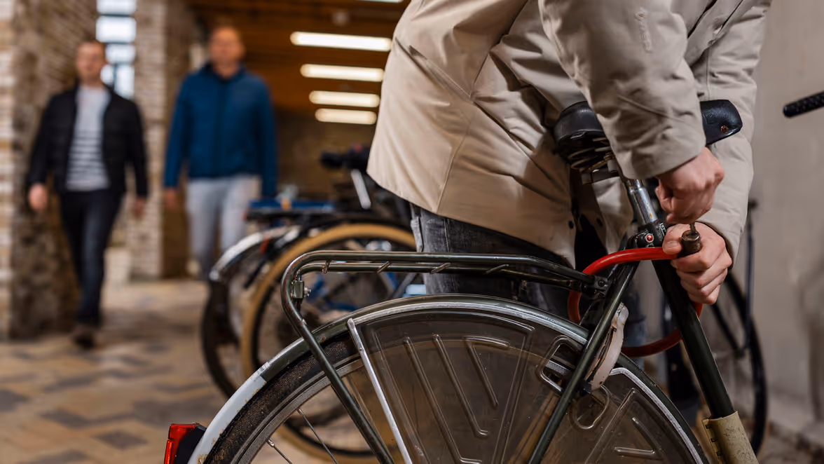 Persoon die een fiets op slot zet in een overdekte fietsenstalling, terwijl op de achtergrond twee mensen komen aanlopen.