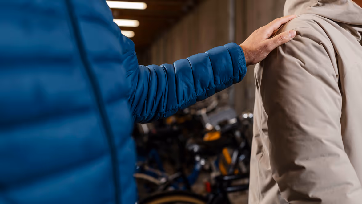 Close-up van een persoon die een hand op de schouder van een potentiele informant legt in een ruimte vol geparkeerde fietsen.
