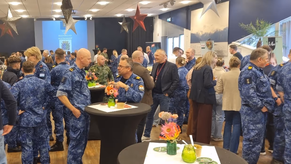 Een groot aantal mensen, militair en burger, in een zaal met ronde tafels.