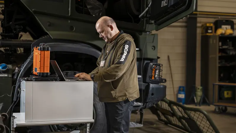 Man kijkt op laptop en staat naast transportvoertuig in werkplaats.
