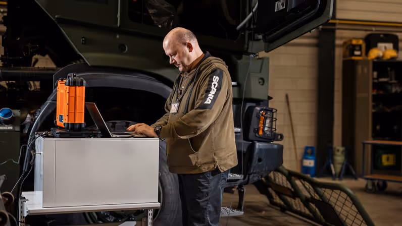 Man kijkt op laptop en staat naast transportvoertuig in werkplaats.