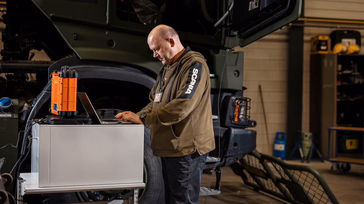 Man kijkt op laptop en staat naast transportvoertuig in werkplaats.